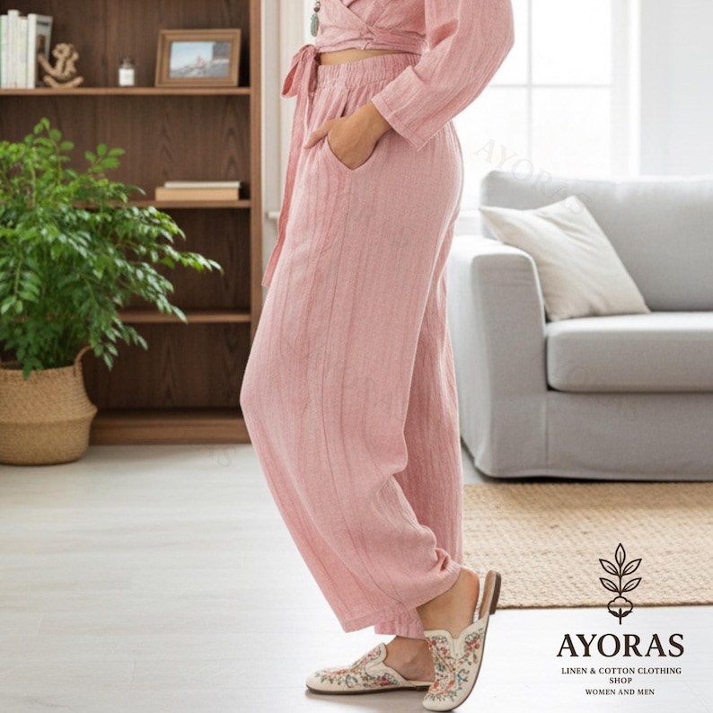 May include: A light pink linen outfit, including a wrap top and wide-leg pants. The outfit has a relaxed fit and features pockets. The person is wearing floral embroidered slip-on shoes. The image is taken indoors, with a couch and bookshelf in the background.