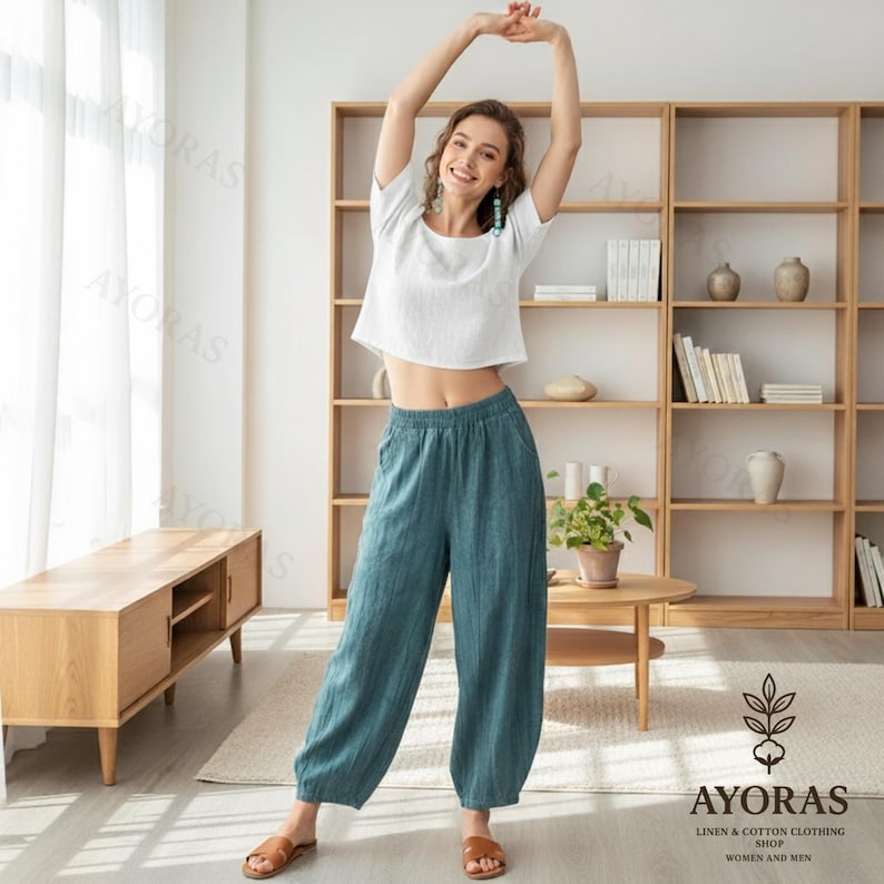 May include: A woman wearing a white crop top and teal linen pants. The pants have an elastic waistband and a relaxed fit. The outfit is casual and comfortable, suitable for warm weather. The image is taken indoors, with a wooden bookshelf in the background.