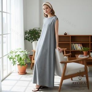 May include: A sleeveless, floor-length gray dress. The dress has a simple, round neckline and a relaxed fit. The image also shows a woman wearing the dress, a white headband, and black sandals. The background includes a window, plants, and wooden furniture.