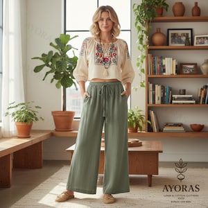 Può includere: Una donna indossa un top color crema ricamato e pantaloni a gamba larga verde oliva. Il top presenta ricami floreali colorati intorno allo scollo e alle maniche. I pantaloni hanno una vita con coulisse. L'immagine è scattata in interni con un tavolo di legno e una libreria sullo sfondo.