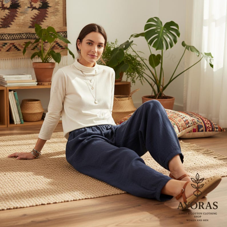 Puede incluir: Una mujer con un cuello alto crema y pantalones azul marino sentada en una alfombra tejida. Lleva un collar y una pulsera plateados. La imagen es de una tienda de ropa de lino y algod&oacute;n, con la marca "AYORAS" visible.