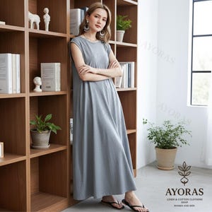 May include: A gray, sleeveless maxi dress with a round neckline. The dress is made of a soft fabric and falls to the ankles. The image also shows a wooden bookshelf and the text "AYORAS LINEN & COTTON CLOTHING SHOP WOMEN AND MEN."