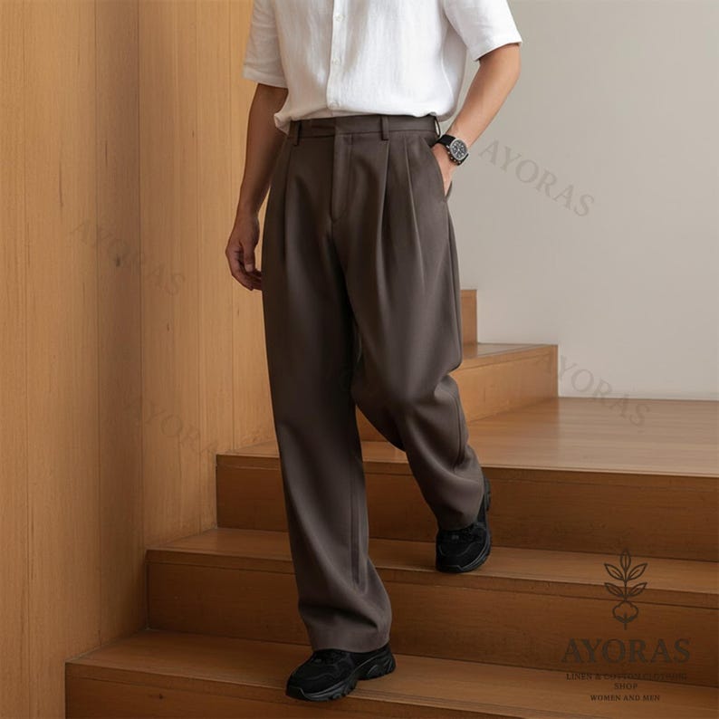 May include: Brown pleated trousers with a relaxed fit. The pants are paired with black sneakers and a white short-sleeve shirt. The trousers have a classic design and are suitable for various occasions.