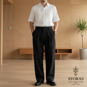 May include: A person wearing a white short-sleeve button-down shirt and black pleated trousers. The outfit is completed with black shoes. The image is taken in a well-lit room with wooden paneling and a plant in the background. The logo for AYORAS is visible.