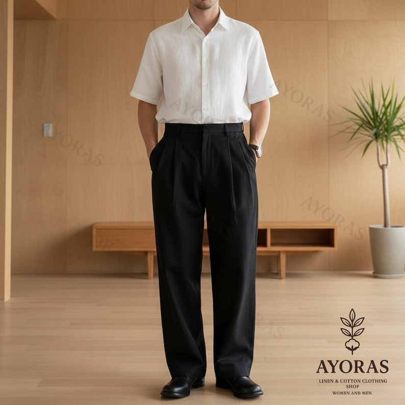 May include: A person wearing a white short-sleeve button-down shirt and black pleated trousers. The outfit is completed with black shoes. The image is taken in a well-lit room with wooden paneling and a plant in the background. The logo for AYORAS is visible.