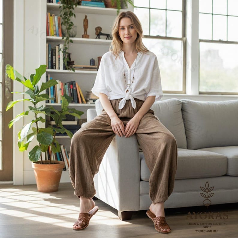 May include: A woman wearing a white linen top tied at the waist and brown linen pants. She is seated on a gray sofa, wearing brown sandals. A bookshelf and a potted plant are in the background. The image is well-lit, showcasing the natural tones of the clothing.
