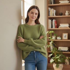 May include: A woman wearing a green crew neck sweater and blue jeans. The sweater has a textured appearance. The woman is standing near a window and a bookshelf. The shop logo "AYORAS" is visible in the image.