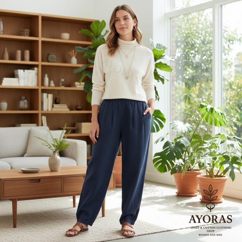 Puede incluir: Una mujer con un jersey de cuello alto color crema y pantalones azul marino. Los pantalones tienen un corte holgado con la parte delantera plisada. El conjunto se completa con sandalias. La imagen est&aacute; tomada en interiores con una estanter&iacute;a de madera y plantas al fondo.
