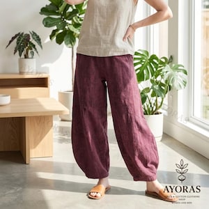 May include: A woman wearing a beige tank top and burgundy linen pants. The pants have a wide, relaxed fit. The image is taken indoors, with natural light and plants in the background. The brand logo is visible in the bottom right corner.