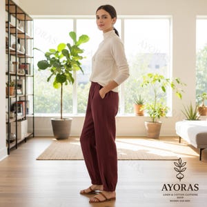 Puede incluir: Una mujer con un jersey de cuello alto crema y pantalones de lino burdeos con tobillo el&aacute;stico. Lleva sandalias marrones. El fondo incluye una estanter&iacute;a, plantas y una ventana. El conjunto es de AYORAS.
