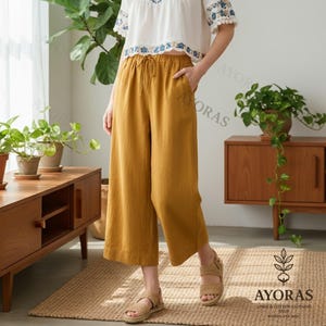 May include: Mustard yellow linen culottes with a drawstring waist. The cropped pants are paired with a white top with blue embroidery. The image also shows woven sandals and a wooden cabinet. The brand logo "AYORAS" is visible.