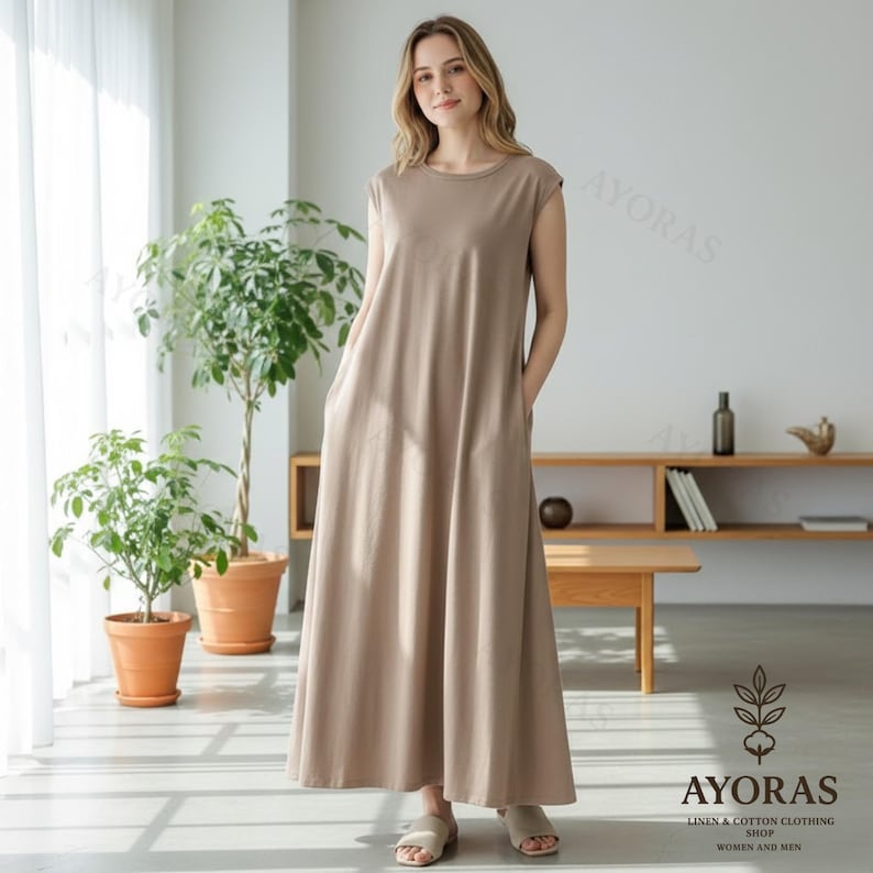 May include: A woman wearing a beige, sleeveless maxi dress with pockets. The dress has a round neckline and a flowing silhouette. The image is taken indoors, with natural light and plants in the background. The dress is from the AYORAS Linen & Cotton Clothing Shop.
