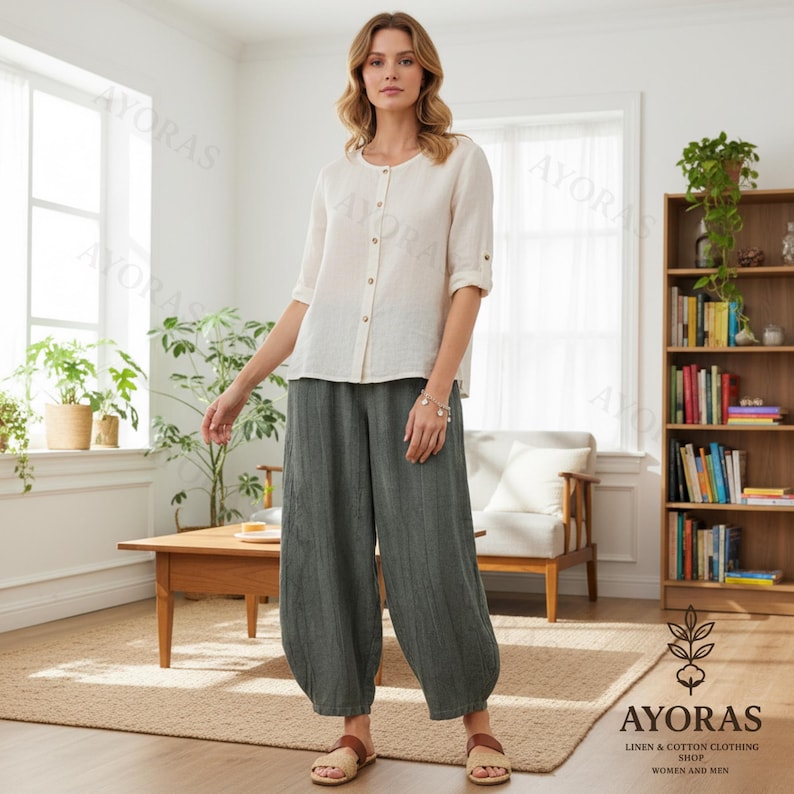 May include: A woman wearing a cream-colored button-down shirt with rolled-up sleeves and olive green, wide-leg pants. The outfit is paired with sandals. The image is taken indoors, with a wooden bookshelf and a coffee table in the background. The brand name "AYORAS" is visible.