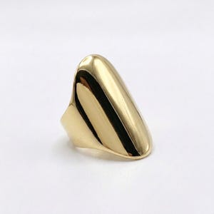 May include: A gold-colored ring with a smooth, elongated design. The ring has a rounded top and a wide band, creating a sleek and modern look. The polished surface reflects light, highlighting its shape.
