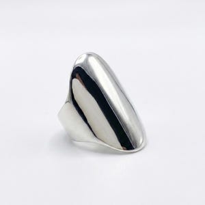 May include: A polished silver ring with a smooth, elongated design. The ring has a rounded top and a wide band, reflecting light. The overall shape is a modern, abstract style, suitable for everyday wear.