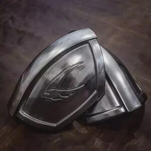 May include: A pair of metallic silver shoulder pauldrons featuring a curved, segmented design with a raised, stylized wing or bird emblem embossed on the front surface. The armor pieces have a polished, reflective finish and a smooth, rounded contour.