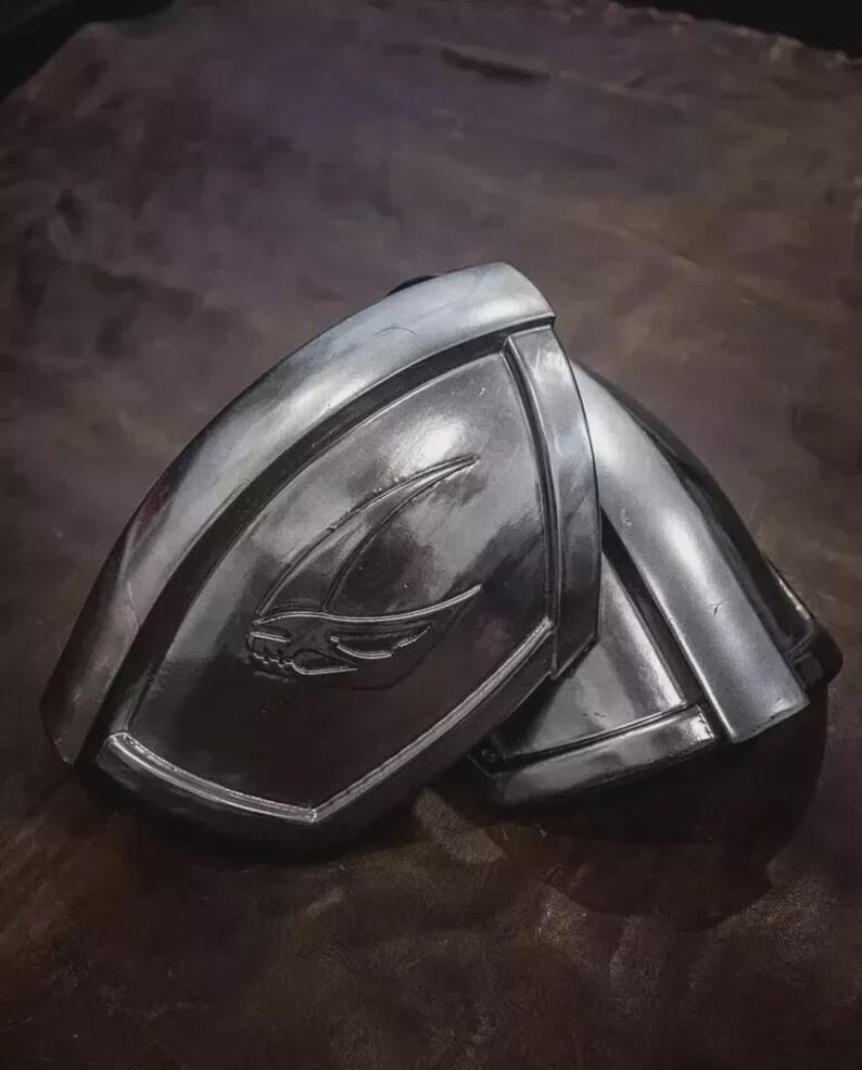 May include: A pair of metallic silver shoulder pauldrons featuring a curved, segmented design with a raised, stylized wing or bird emblem embossed on the front surface. The armor pieces have a polished, reflective finish and a smooth, rounded contour.