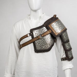 May include: A shoulder and upper arm armour piece featuring silver-toned metal plates with intricate embossed knotwork patterns, secured with brown leather straps, metal buckles, and dark brown leather trim on a white long-sleeved shirt.