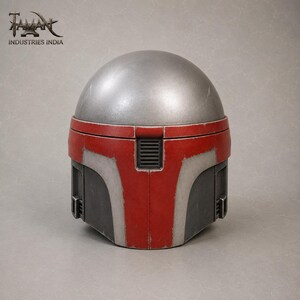 May include: A silver and red Mandalorian helmet with a rounded top and a rectangular visor. The helmet has a red faceplate with silver accents and a black vent. The logo "TAMAN INDUSTRIES INDIA" is visible in the top left corner.