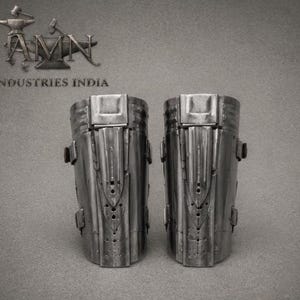 May include: A pair of metallic greaves featuring a polished steel finish with vertical ridges, central decorative plates, and side buckles. The text AMN INDUSTRIES INDIA is visible in the upper left corner with an anvil and hammer logo.