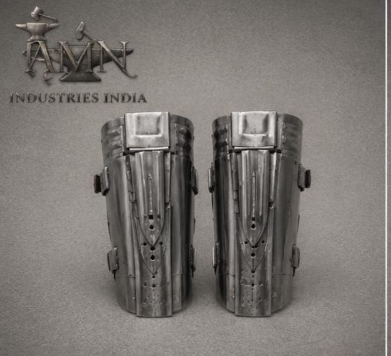 May include: A pair of metallic greaves featuring a polished steel finish with vertical ridges, central decorative plates, and side buckles. The text AMN INDUSTRIES INDIA is visible in the upper left corner with an anvil and hammer logo.