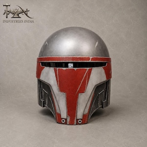 May include: A silver and red helmet with a weathered appearance. The helmet features a rounded top and a faceplate with red and white accents. The text "INDUSTRIES INDIA" is visible in the upper left corner.