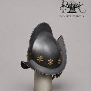 May include: A dark gray metal helmet with a wide brim and gold fleur-de-lis accents. The helmet is displayed on a white cylindrical stand. The logo "INDUSTRIES INDIA" is visible in the background.
