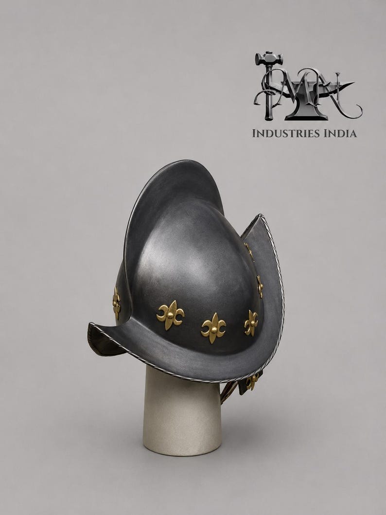 May include: A dark gray metal helmet with a wide brim and gold fleur-de-lis accents. The helmet is displayed on a white cylindrical stand. The logo "INDUSTRIES INDIA" is visible in the background.