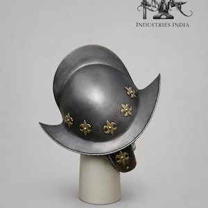 May include: A silver-colored metal helmet with a wide brim and decorative gold-colored fleur-de-lis accents. The helmet is displayed on a white cylindrical stand, showcasing its historical design and craftsmanship. The text "INDUSTRIES INDIA" is visible.
