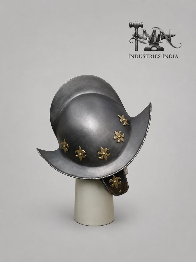 May include: A silver-colored metal helmet with a wide brim and decorative gold-colored fleur-de-lis accents. The helmet is displayed on a white cylindrical stand, showcasing its historical design and craftsmanship. The text "INDUSTRIES INDIA" is visible.