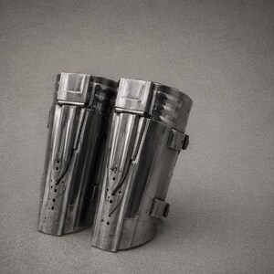 May include: A pair of metallic forearm guards featuring a polished dark grey finish, vertical raised panels, and small circular perforations along the lower section. Each piece includes a top rectangular clasp and side straps for secure fastening.