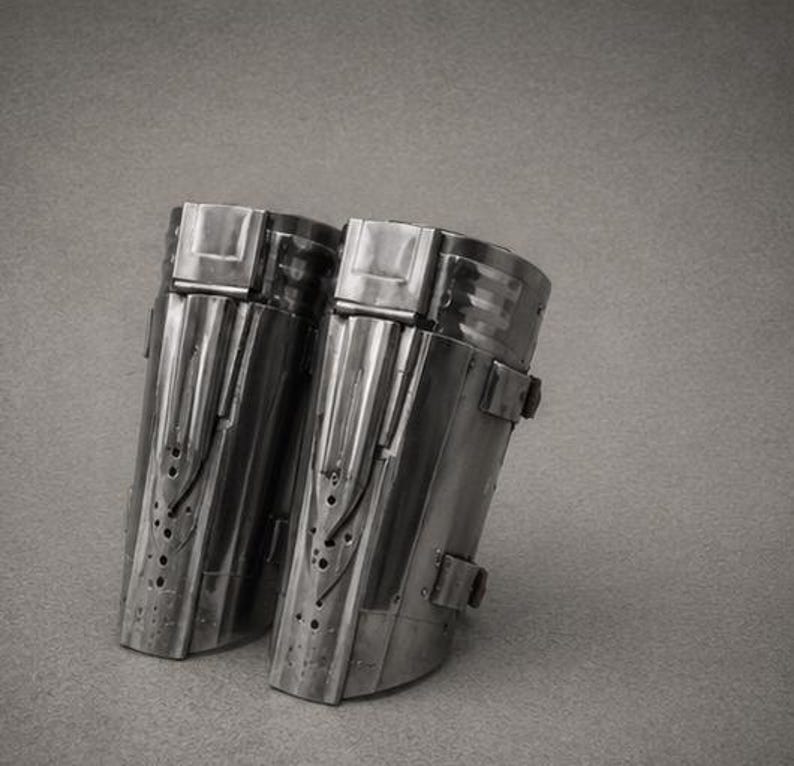 May include: A pair of metallic forearm guards featuring a polished dark grey finish, vertical raised panels, and small circular perforations along the lower section. Each piece includes a top rectangular clasp and side straps for secure fastening.