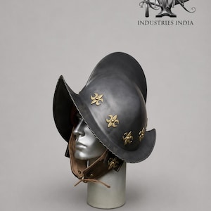 May include: A dark gray metal helmet with a wide brim and decorative gold fleur-de-lis emblems. The helmet is displayed on a mannequin head with a brown leather neck guard. The text "TMN INDUSTRIES INDIA" is visible in the upper right corner.