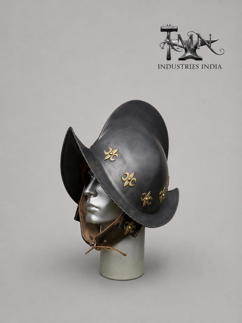 May include: A dark gray metal helmet with a wide brim and decorative gold fleur-de-lis emblems. The helmet is displayed on a mannequin head with a brown leather neck guard. The text "TMN INDUSTRIES INDIA" is visible in the upper right corner.