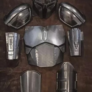 May include: A set of metallic silver armor pieces including a full-face helmet, two shoulder pauldrons, a chest plate with a central emblem, two forearm gauntlets with detailed mechanical textures, and two lower leg greaves arranged on a dark surface.