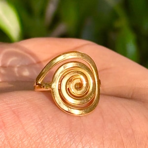 May include: A gold-colored ring with a spiral design. The ring features a central spiral surrounded by a curved band, creating a unique and eye-catching shape. The ring is shown on a hand, with a blurred green background.