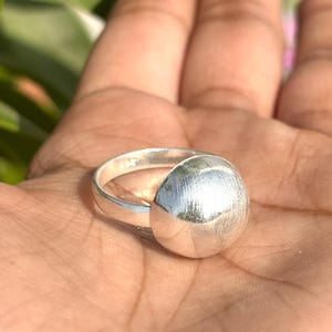 Puede incluir: Un anillo de plata con un diseño grande y texturizado de media esfera. El anillo está engarzado en una banda sencilla y tiene un acabado de metal cepillado. El anillo se muestra en la mano de una persona.