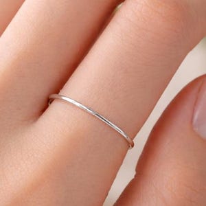 May include: A slender silver ring is displayed on a finger. The ring is a thin, simple band, catching the light. The image highlights the jewellery, emphasizing its minimalist design and elegant simplicity. The ring's slim profile suggests a refined aesthetic.