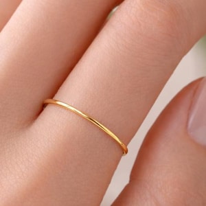 Puede incluir: Un delicado y fino anillo de oro en un dedo. El anillo es una banda simple, que muestra un diseño minimalista. La superficie pulida del anillo refleja la luz, resaltando su textura suave. Una pieza de joyería clásica.