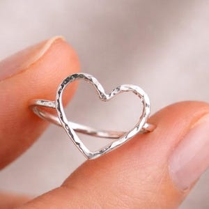 May include: A silver ring with a heart-shaped outline. The ring is held up, displaying the open heart design. The metal has a textured finish, adding detail to the simple, elegant jewellery. The ring's design is a classic.
