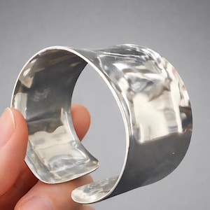 Könnte beinhalten: Ein breites, offenes Silberarmband mit gehammerter Textur. Das Armband wird in einer Hand vor grauem Hintergrund gehalten. Das Armband ist etwa 5 cm breit.