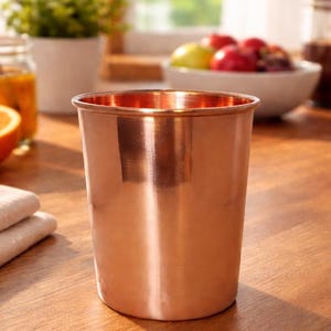 以下が含まれることがあります： 滑らかで反射する表面を持つ銅色のグラス。グラスは円筒形で、縁がわずかに広がっています。背景には木製のテーブル、半分に切ったオレンジ、オレンジ色の液体が入った瓶が含まれています。