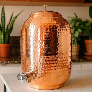 May include: A hammered copper water dispenser with a spigot. The dispenser has a lid with a small handle. The copper has a shiny, reflective surface. The dispenser is sitting on a white countertop.