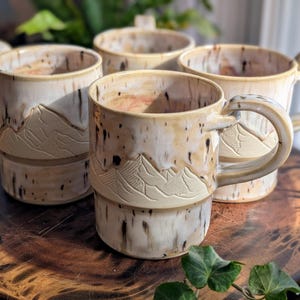 May include: Four ceramic mugs with mountain designs. The mugs are beige with brown speckles and feature a mountain range carved into the side. Each mug has a large, curved handle. The mugs are arranged on a wooden surface.