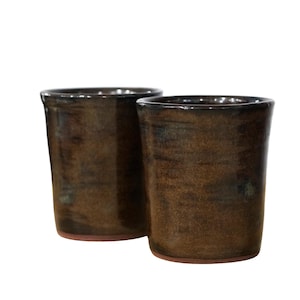 Peut inclure: Deux tasses cylindriques avec une finition émaillée brun foncé brillante. La surface extérieure présente des variations subtiles de tons et de textures, tandis que la base de chaque tasse révèle un bord en argile brun rougeâtre naturel.