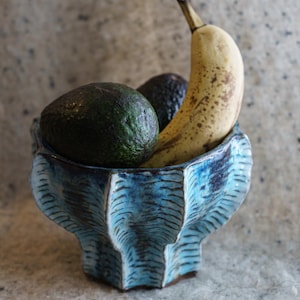 Puede incluir: Un cuenco de cerámica azul con exterior texturizado contiene dos aguacates y un plátano. El plátano es amarillo con manchas marrones. El cuenco tiene una forma única y es de color azul vibrante.