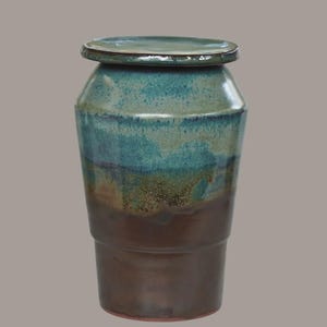 Peut inclure: Un pot en céramique avec couvercle, doté d'un corps cylindrique effilé. La partie supérieure est recouverte d'une glaçure bleu sarcelle vive, qui se fond dans une teinte brune terreuse profonde sur la section inférieure. Finition lisse.