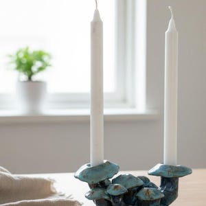 May include: A ceramic candle holder featuring a cluster of blue mushroom shapes, holding two white taper candles. The candle holder is on a wooden surface, with a window and a small potted plant in the background.