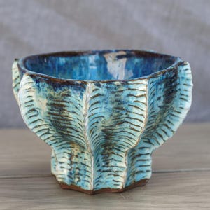 May include: A ceramic bowl with a fluted, scalloped exterior featuring a vibrant blue and turquoise glaze. The surface has a textured, ribbed pattern with dark brown accents in the crevices, and the interior is finished in a deep, glossy blue.