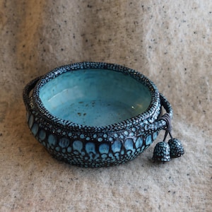 Może przedstawiać: Ręcznie wykonana ceramiczna miska z turkusowym wnętrzem i teksturowanym, ciemnoniebieskim zewnętrzem. Miska posiada dwa uchwyty i ozdobne frędzle. Zewnętrzna strona ma wzór z małych kropek i większych, owalnych kształtów.